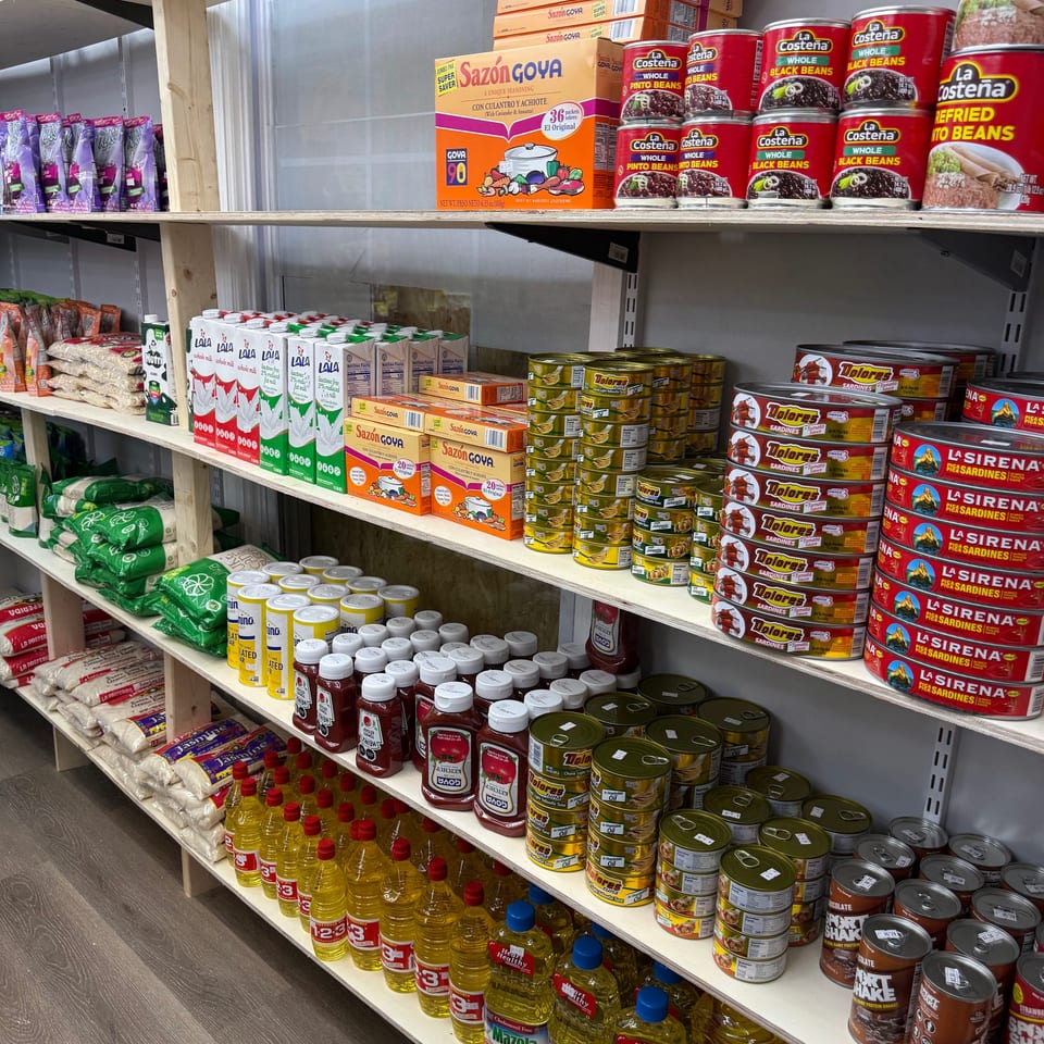 Grocery store shelves stocked with Latin American canned beans, sardines, seasoning, milk, rice, cooking oil, and other packaged foods.